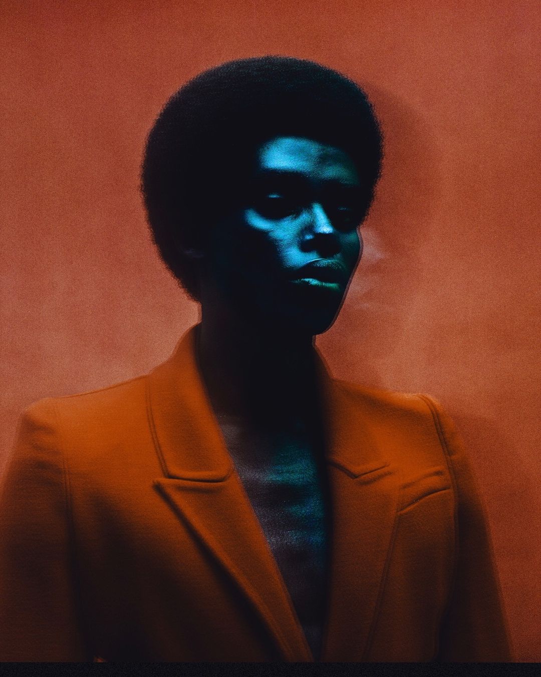 Portrait of a Black female model with an afro hairstyle, wearing an orange jacket, and standing against a textured orange background.