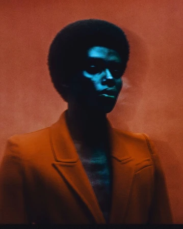 Portrait of a black man with an afro wearing a structured orange blazer, illuminated by striking blue light against a textured background