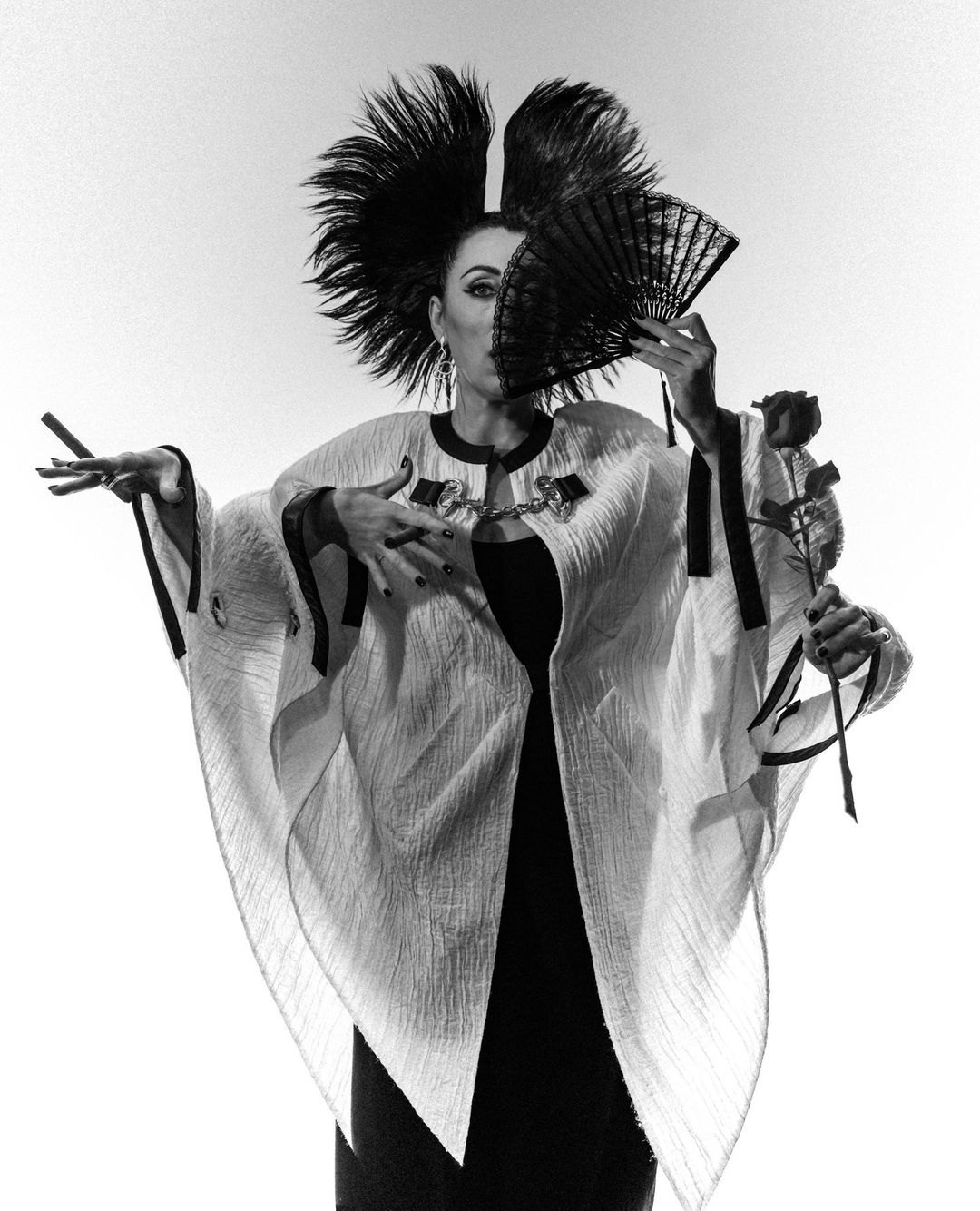 Black and white portrait of a woman with an elaborate feather headpiece, holding a fan and a rose against a plain background.