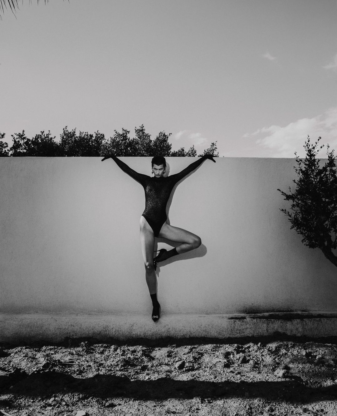 Monochrome full-length portrait of a dancer in a black bodysuit posing gracefully in front of an outdoor concrete wall.