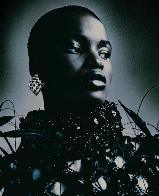 Close-up of a Black woman in elegant beaded attire and diamond earrings against a dark gray backdrop, facing slightly right.
