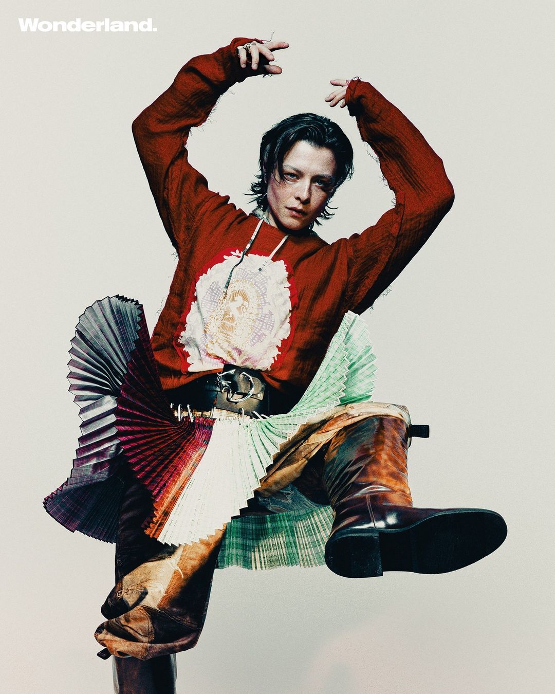A model with dark hair poses in a red sweater, pleated skirt and high-heeled boots against a light background for a studio portrait.