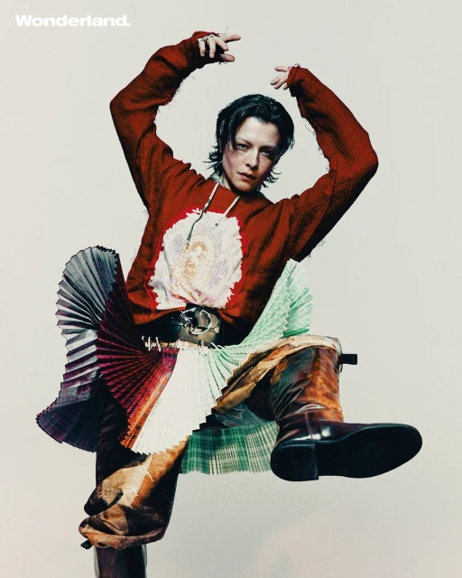 A model with dark hair poses in a red sweater, pleated skirt and high-heeled boots against a light background for a studio portrait.