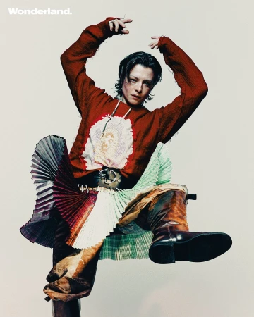 A model with dark hair poses in a red sweater, pleated skirt and high-heeled boots against a light background for a studio portrait.