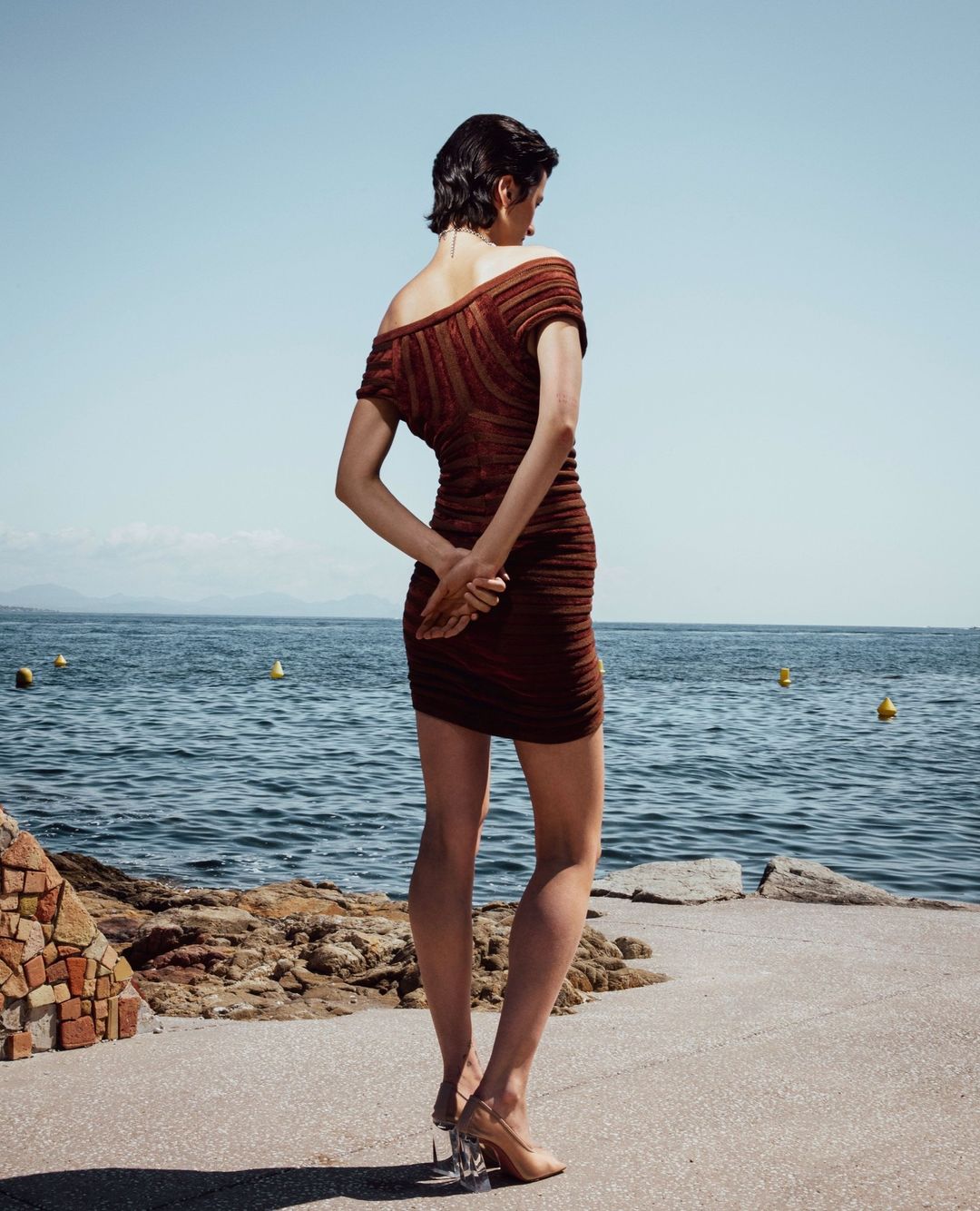Woman in a brown ruched mini dress with tan heels stands on a concrete walkway by the sea on a clear sunny day. Fashion shot.