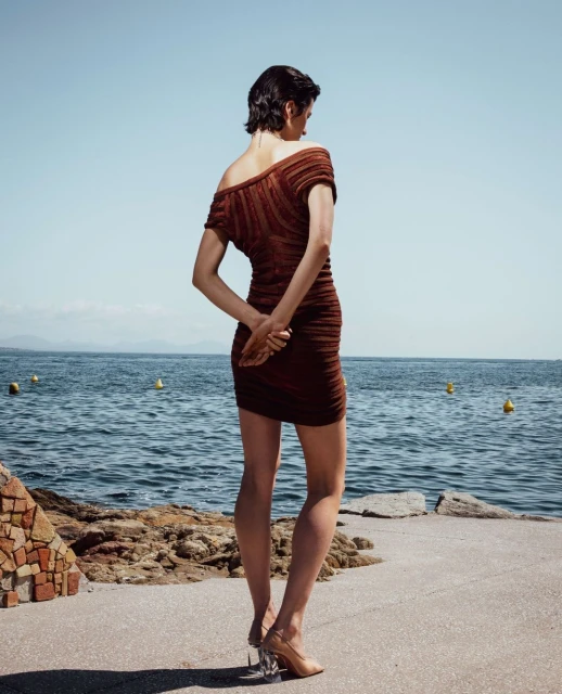 Woman in a brown ruched mini dress with tan heels stands on a concrete walkway by the sea on a clear sunny day. Fashion shot.