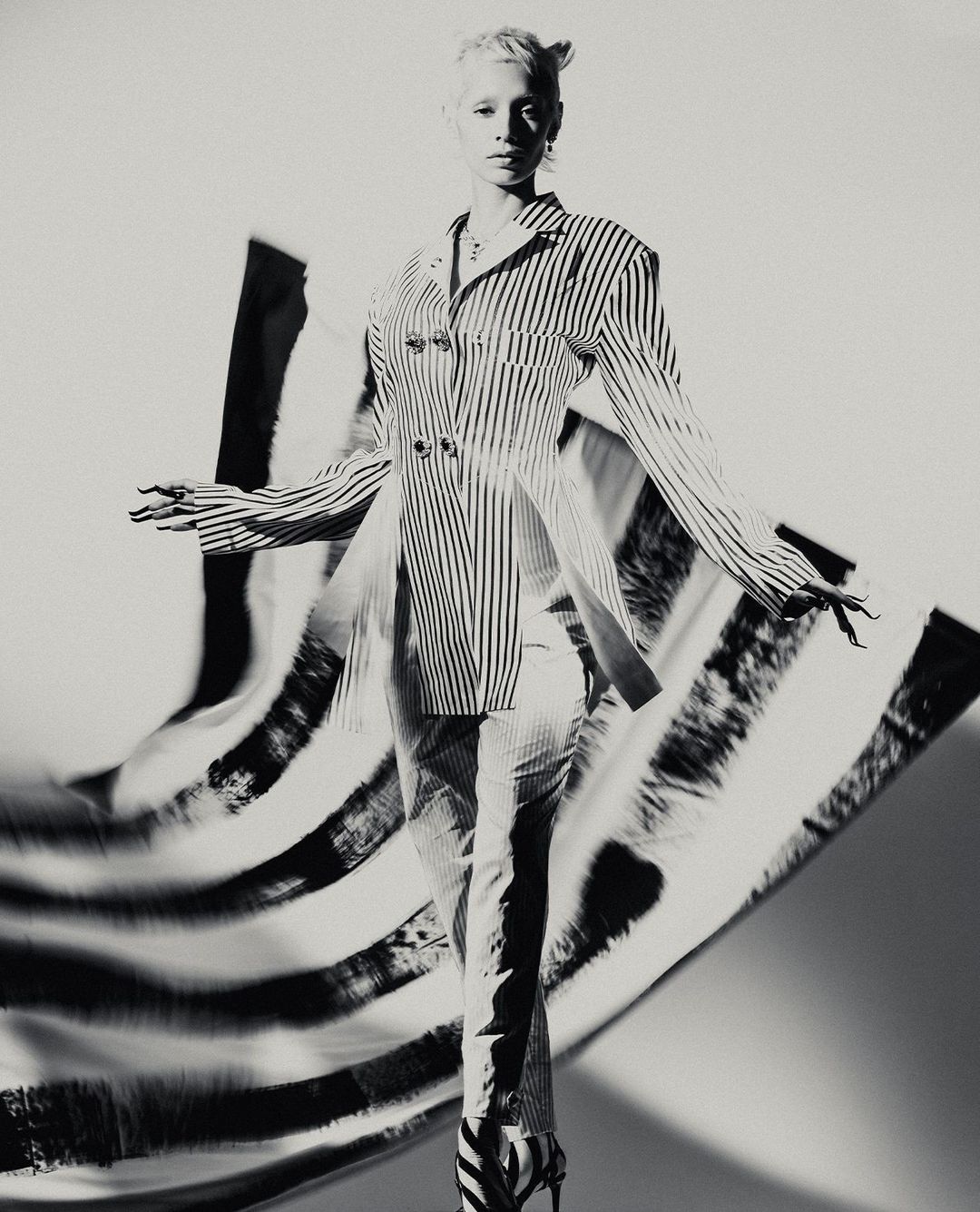 Black-and-white fashion shot of a slender adult woman wearing a striped jacket and pants, striking a pose against a blurred background.