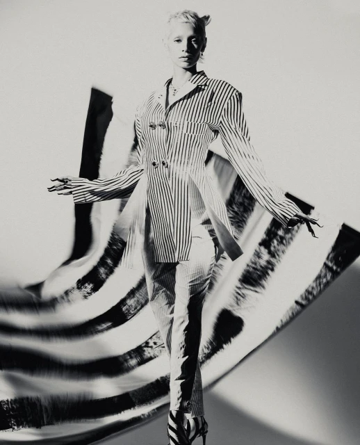 Black-and-white fashion shot of a slender adult woman wearing a striped jacket and pants, striking a pose against a blurred background.