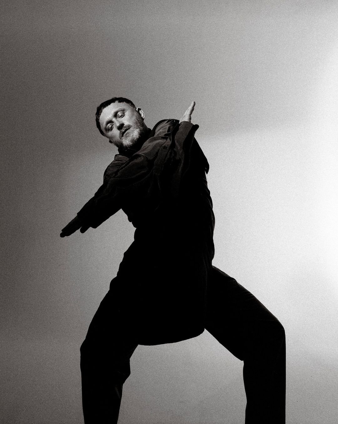 Monochromatic portrait of a bearded adult male in wide-legged stance, wearing dark clothing and extending his arms against a light background.