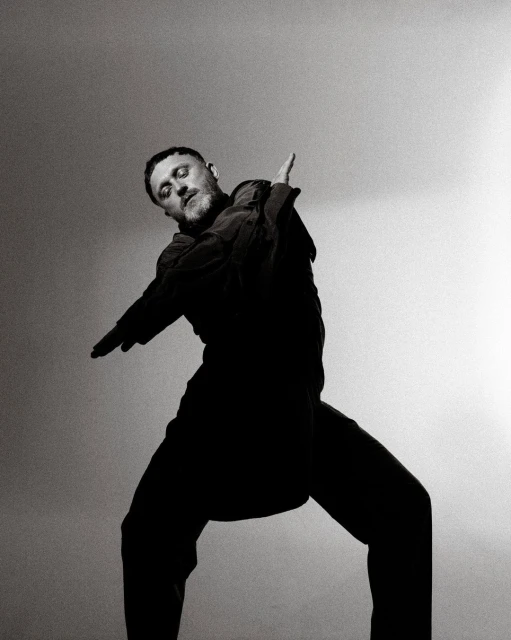Monochromatic portrait of a bearded adult male in wide-legged stance, wearing dark clothing and extending his arms against a light background.