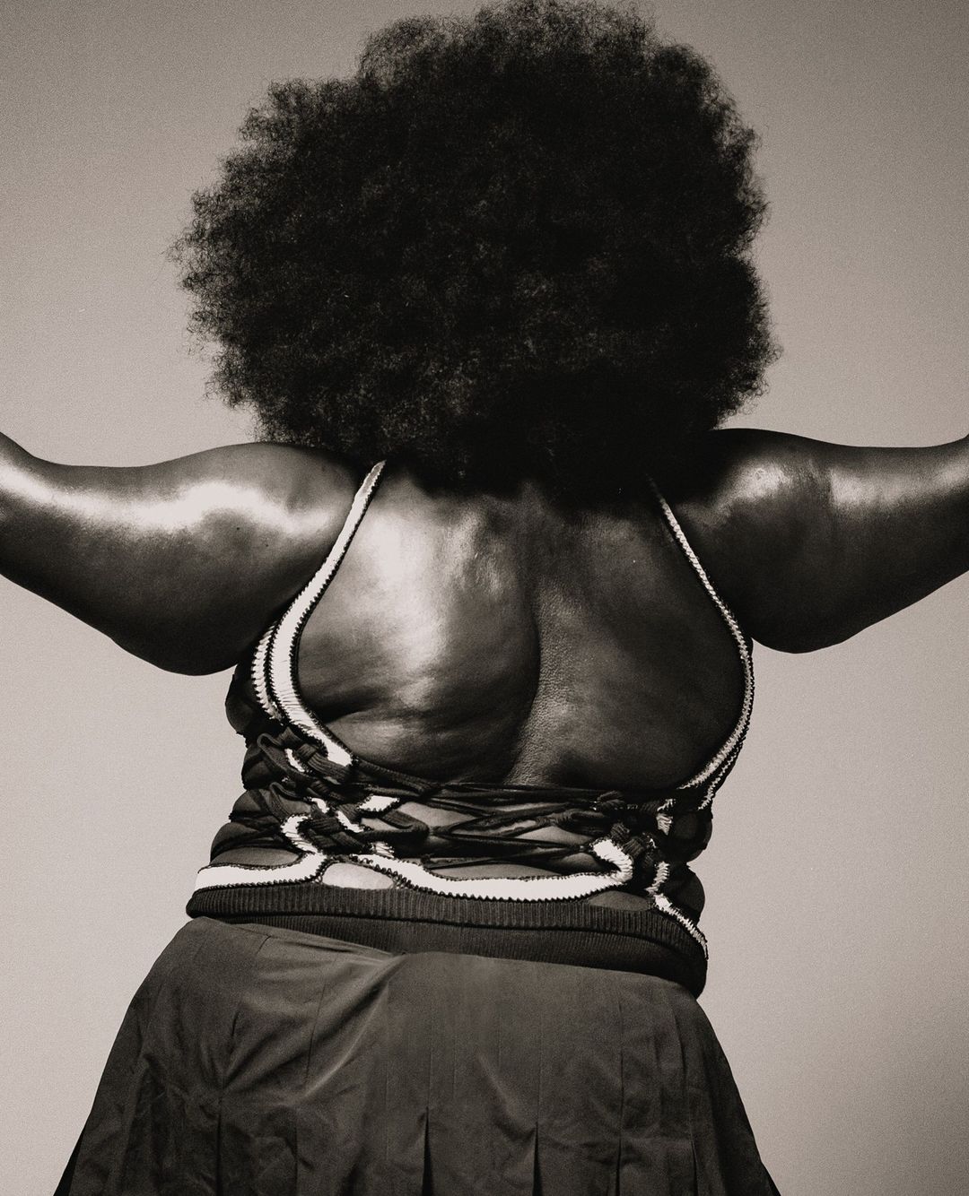 Back view of an adult woman with an afro hairstyle, wearing a dark skirt and a knitted top with a lace-up back, in a studio shot.