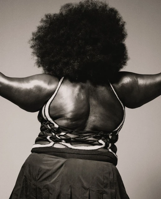 Back view of an adult woman with an afro hairstyle, wearing a dark skirt and a knitted top with a lace-up back, in a studio shot.