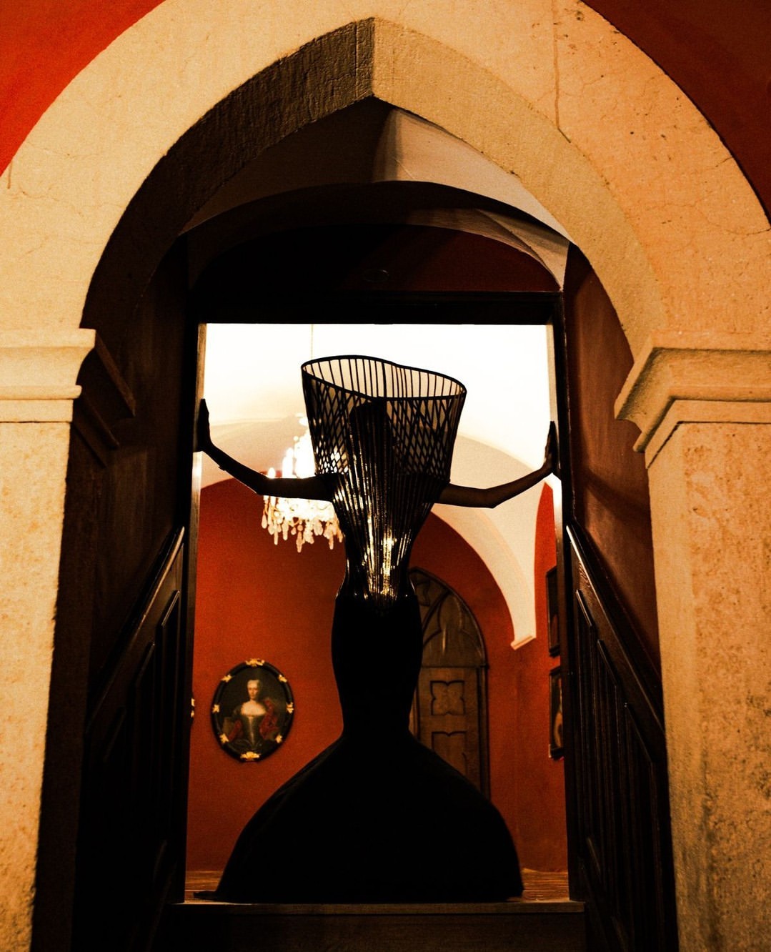 Architectural indoor shot of an abstract, full-length wire sculpture of a woman in a dress near a stone archway and stairway.