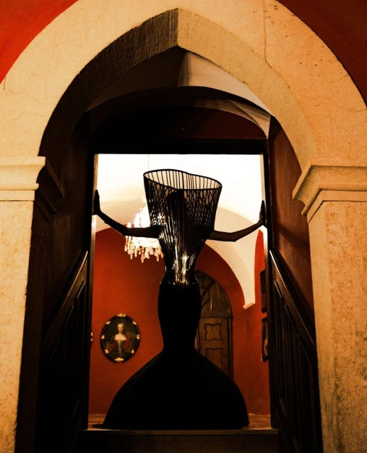 Architectural indoor shot of an abstract, full-length wire sculpture of a woman in a dress near a stone archway and stairway.