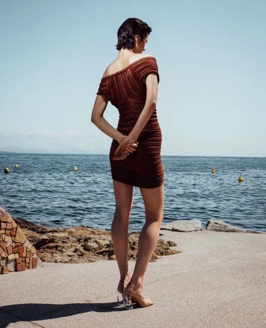 A slender woman with short dark hair stands on a concrete platform with her back turned, facing the ocean on a sunny day.