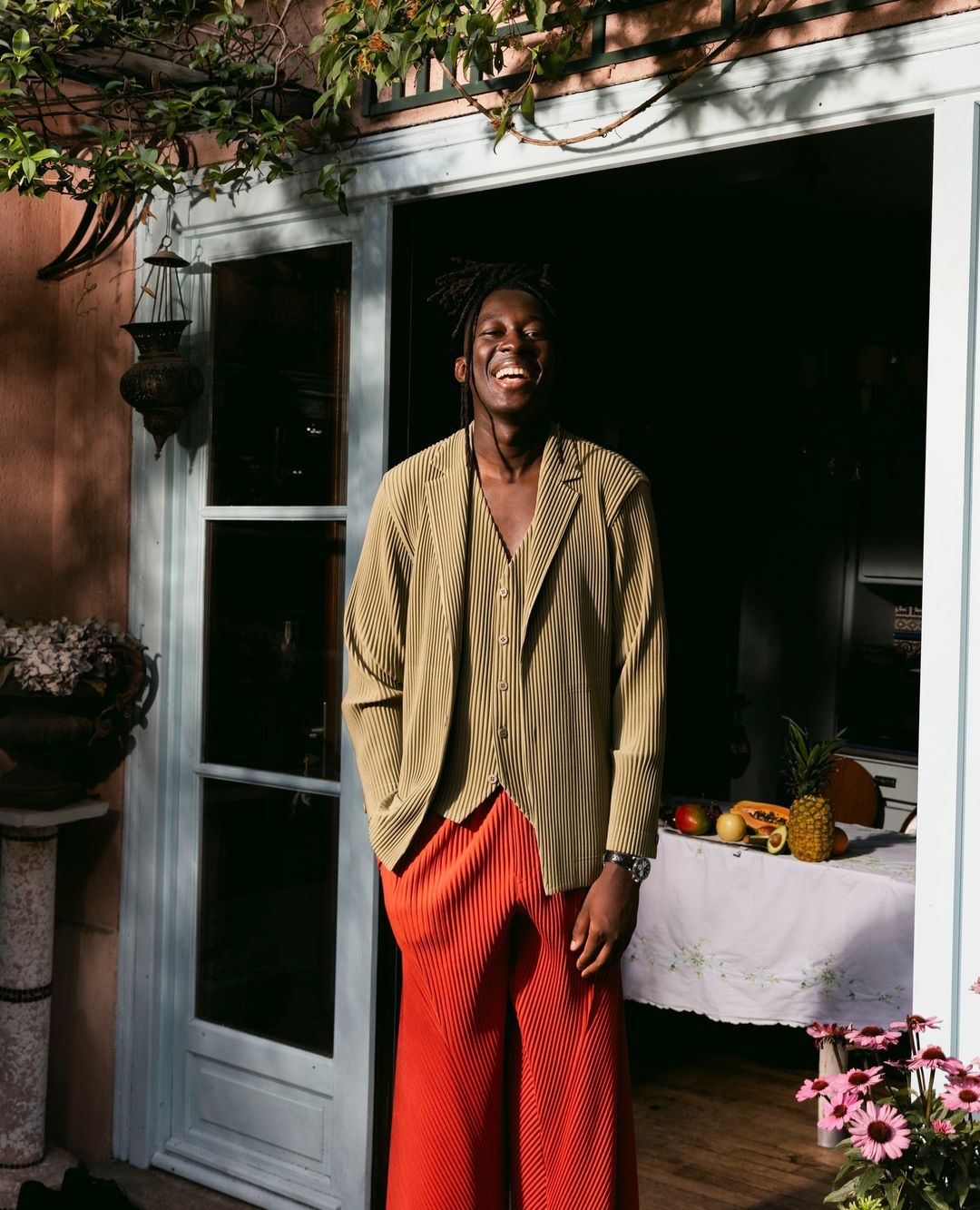Adult male smiles in front of open door wearing an olive jacket, vest, and orange pants, next to a table with assorted fruit.