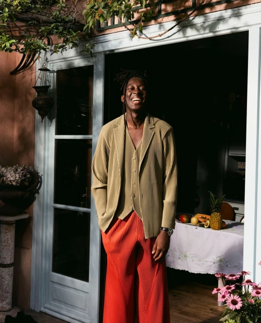 Adult male smiles in front of open door wearing an olive jacket, vest, and orange pants, next to a table with assorted fruit.