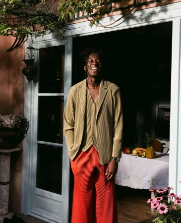 Adult male smiles in front of open door wearing an olive jacket, vest, and orange pants, next to a table with assorted fruit.