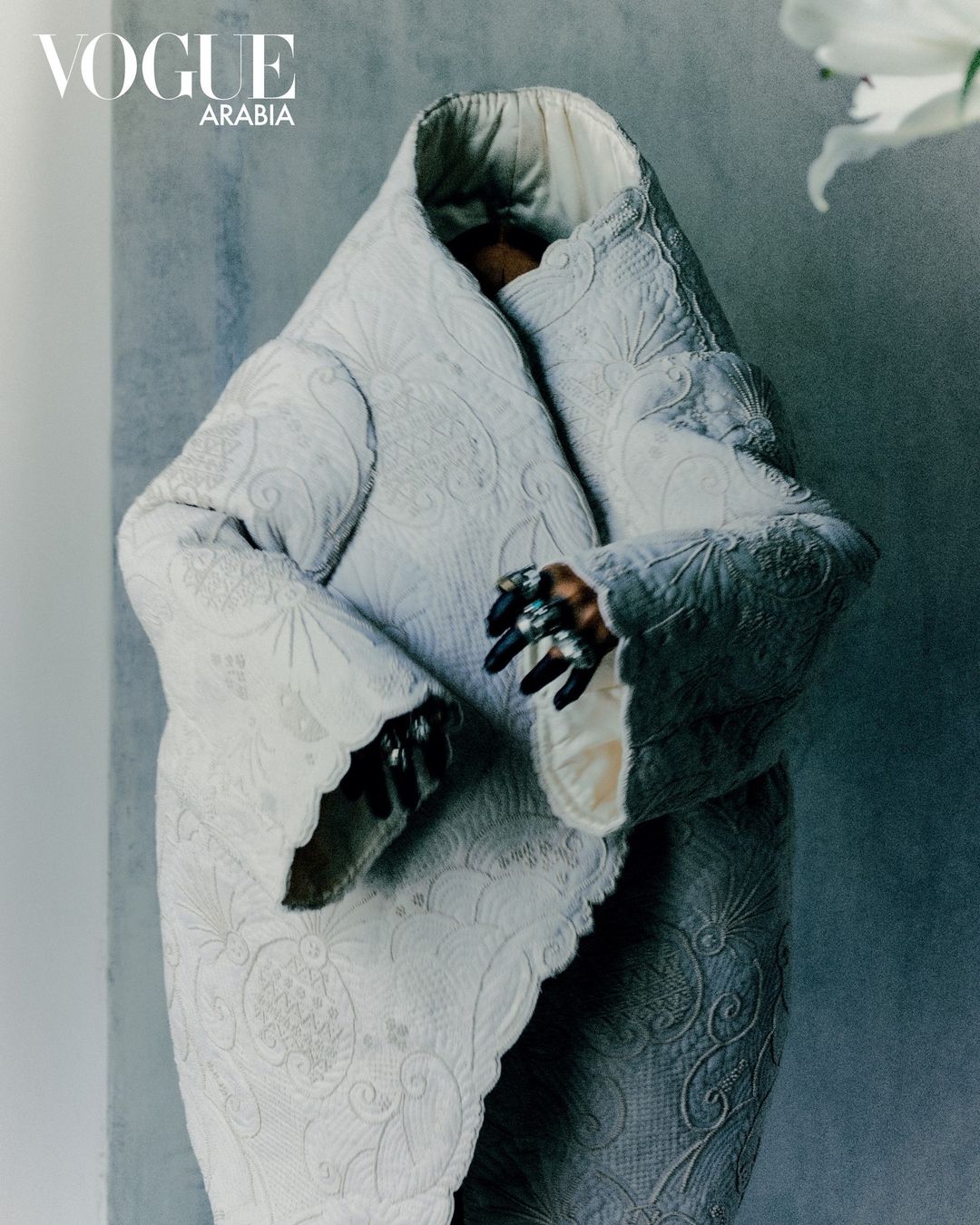 Avant-garde fashion shot with a figure in an elaborate white coat, ornate ring, against a textured grey background and white lily.