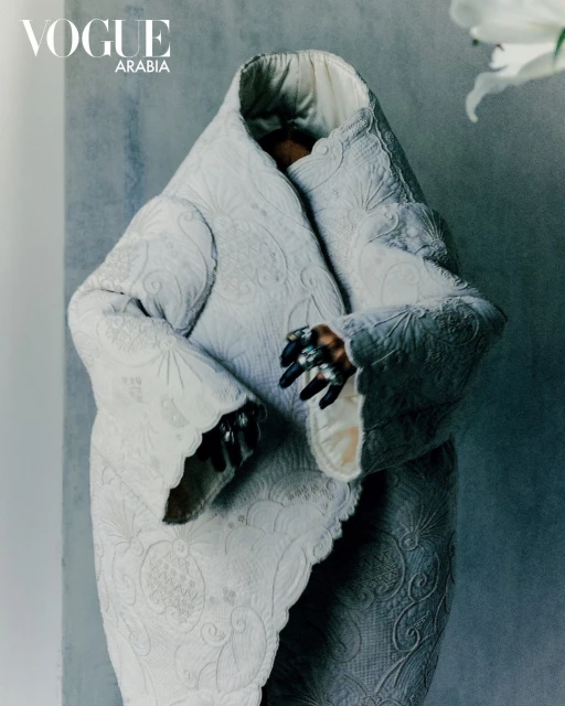 Avant-garde fashion shot with a figure in an elaborate white coat, ornate ring, against a textured grey background and white lily.