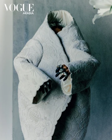 Avant-garde fashion shot with a figure in an elaborate white coat, ornate ring, against a textured grey background and white lily.