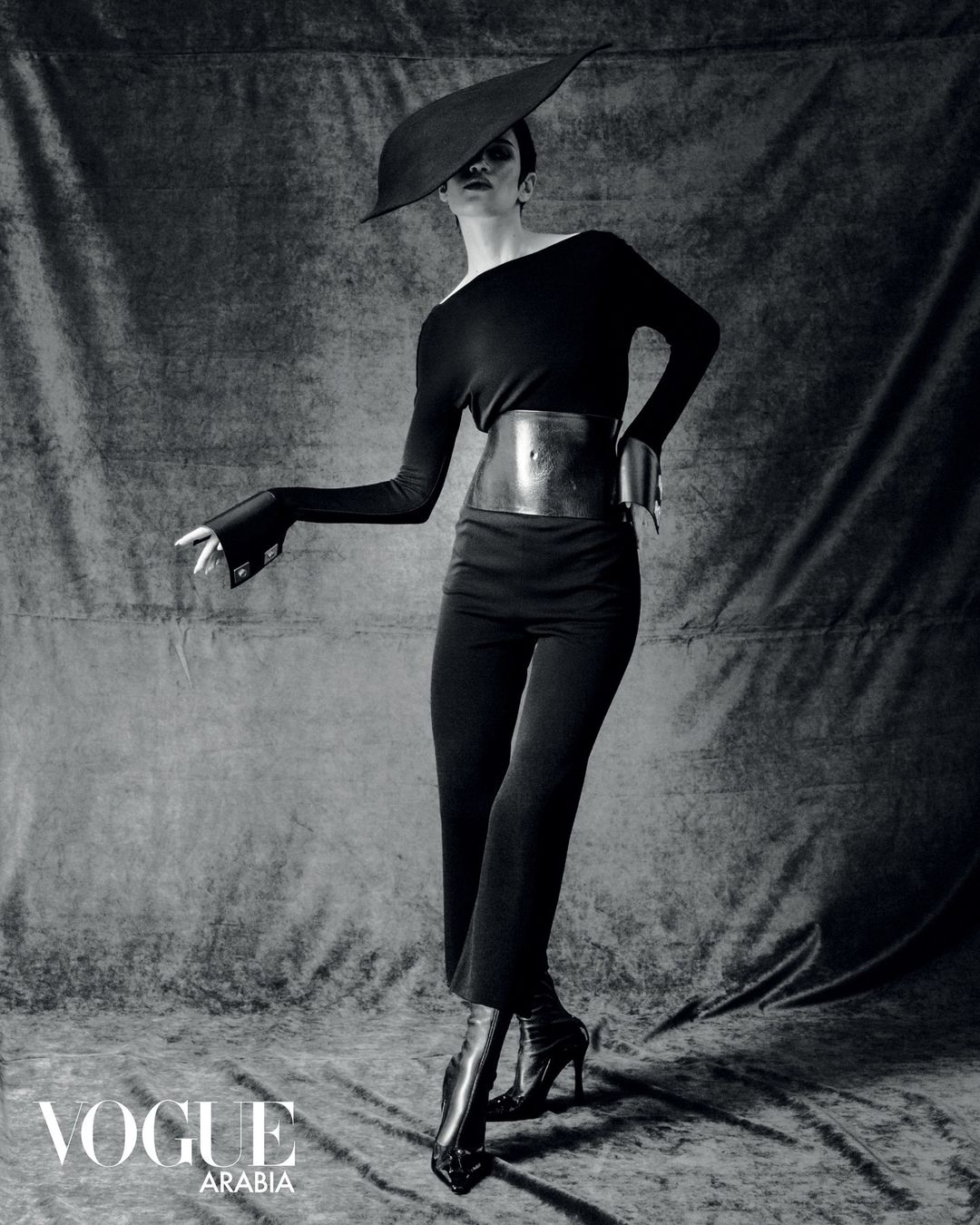 Elegant fashion shot of a woman in a black dress, wide belt, high heels, and hat, posing against a textured grey backdrop.