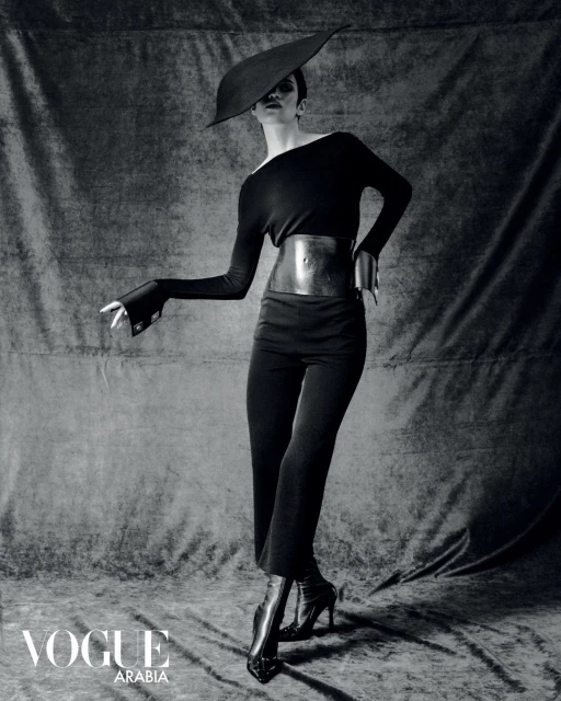 Elegant fashion shot of a woman in a black dress, wide belt, high heels, and hat, posing against a textured grey backdrop.