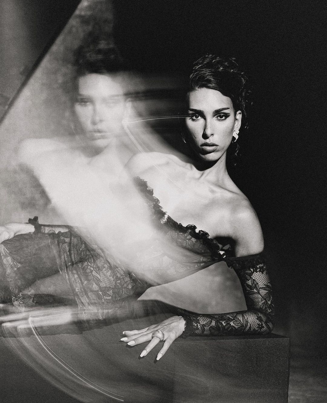 Monochrome studio portrait of an adult woman with lace sleeves posing against a dark background, reflecting a ghosted image.