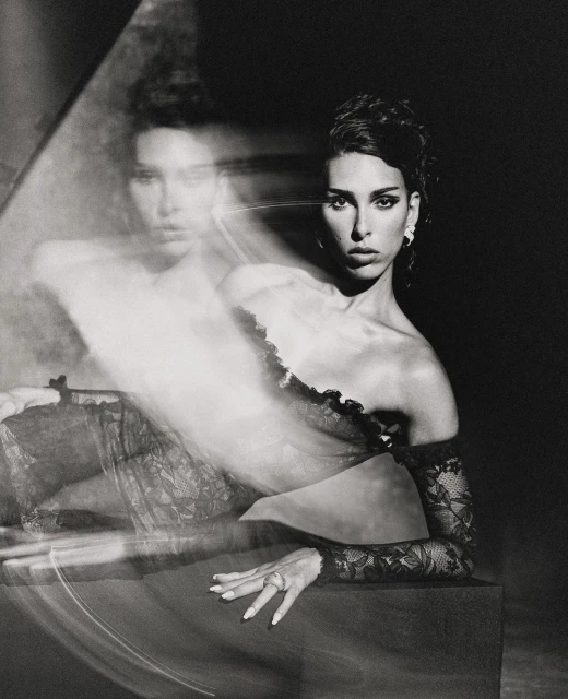 Monochrome studio portrait of an adult woman with lace sleeves posing against a dark background, reflecting a ghosted image.