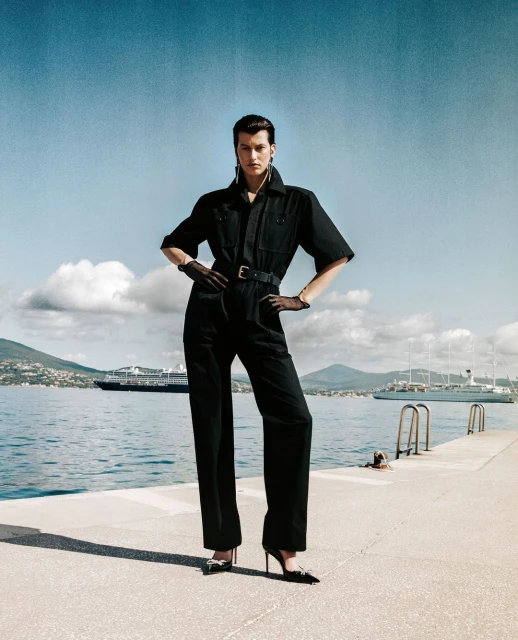 Fashion shot of a woman in a black jumpsuit with a belt and high heels, posing on a pier with boats on the water and mountains in the distance.