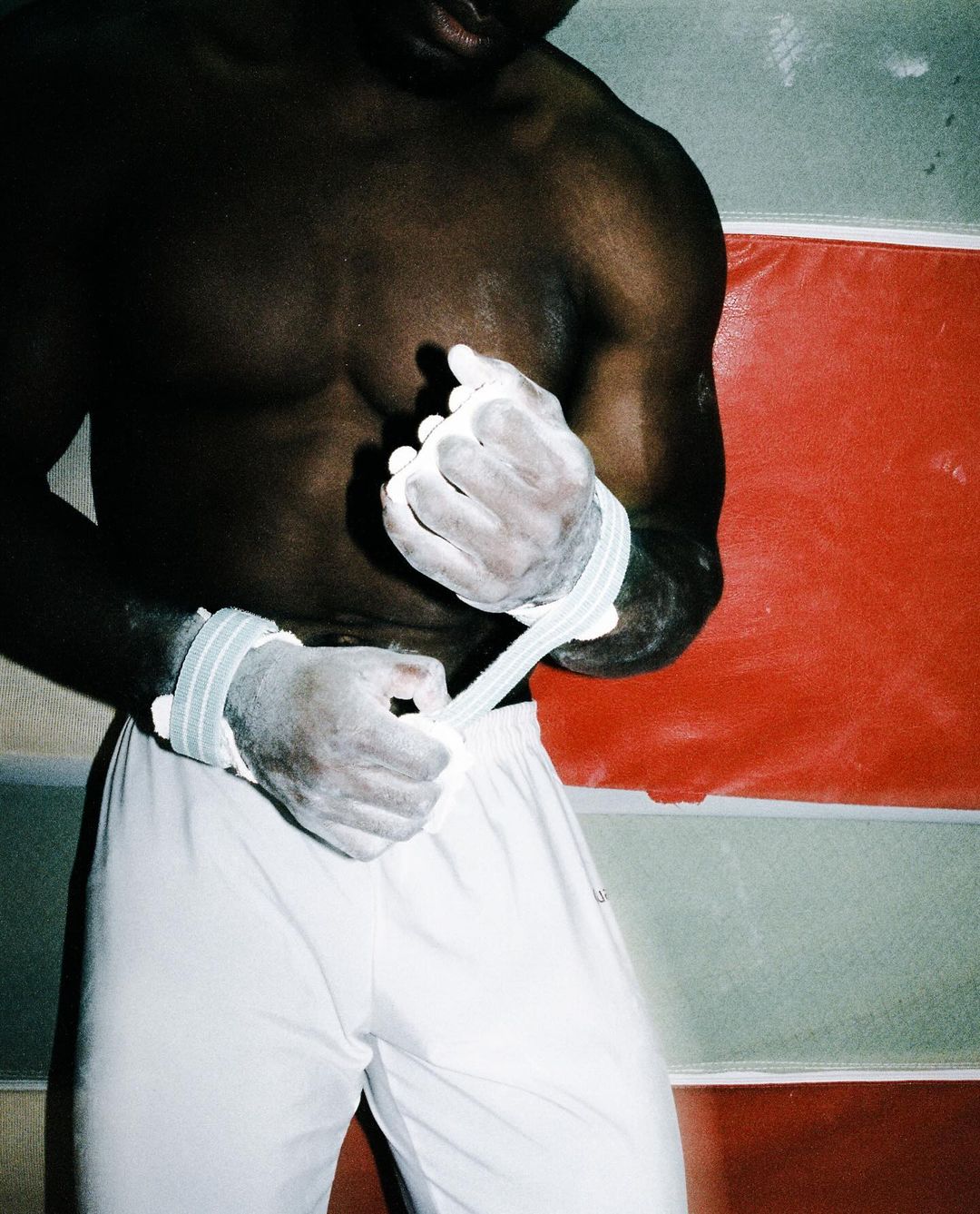 Dark-skinned athlete prepares for gymnastics, secures hand wraps with chalk powder, wearing white athletic pants in an indoor space.
