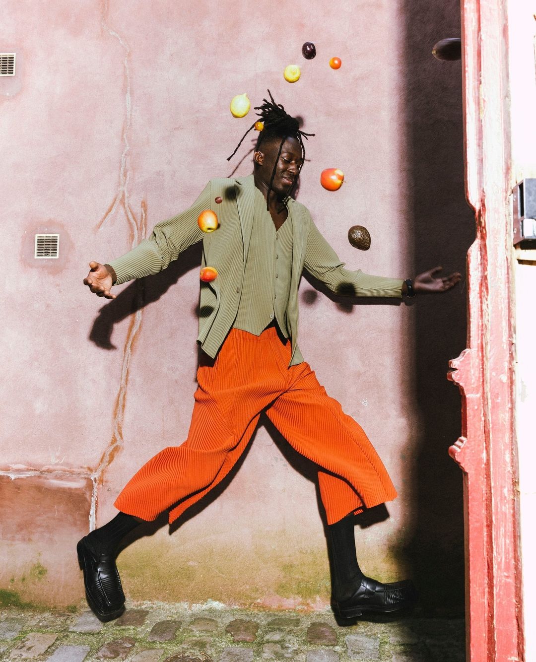 A Black man in a green suit and orange pants jumps with fruit floating around him in front of a textured, pink building exterior.