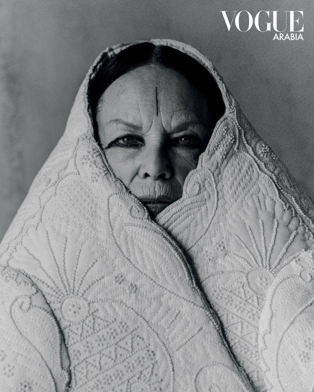 Black and white portrait of an elderly woman wrapped in a patterned white shawl, styled for Vogue Arabia, against a neutral background.