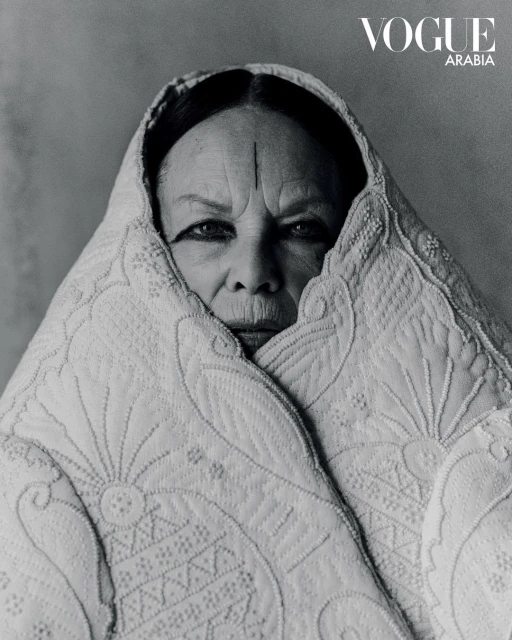 Black and white portrait of an elderly woman wrapped in a patterned white shawl, styled for Vogue Arabia, against a neutral background.