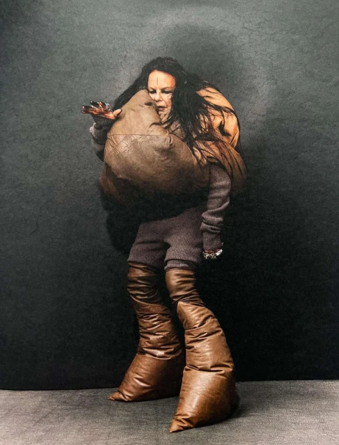 A woman with long dark hair and unusual brown leather fashion, including baggy boots and a sculptural shoulder bag, in a studio.