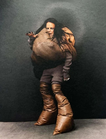 A woman with long dark hair and unusual brown leather fashion, including baggy boots and a sculptural shoulder bag, in a studio.