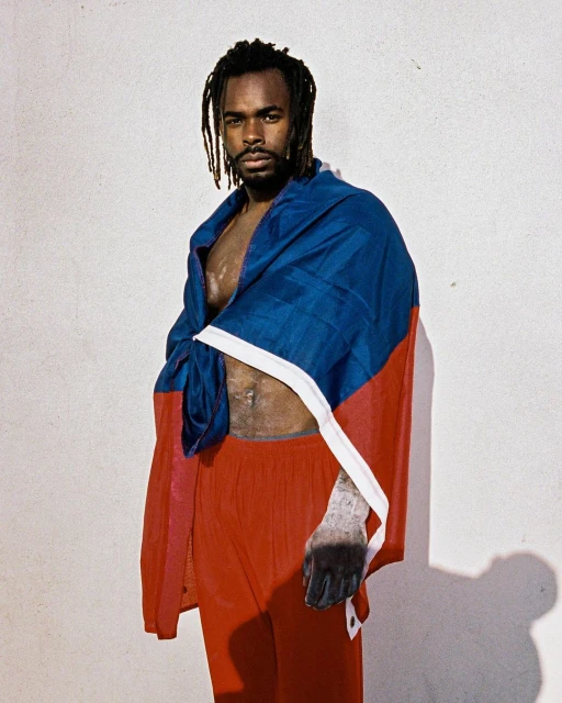 Portrait of a Black adult male wearing a blue, white, and red flag draped over his shoulders in a neutral indoor environment.