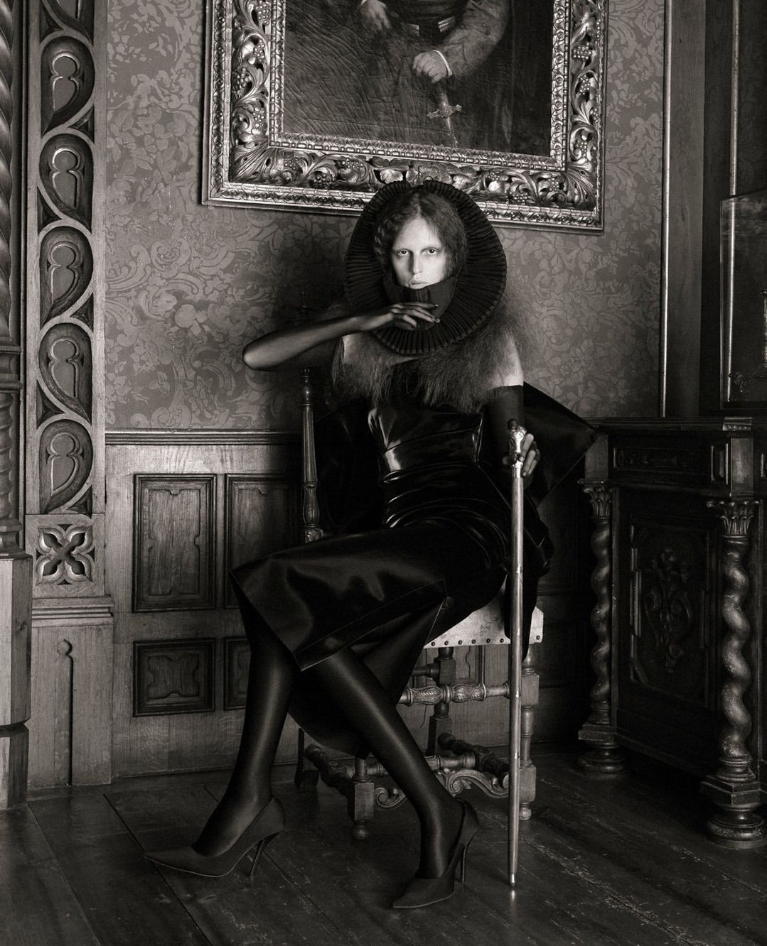 A woman seated on a chair in an elegant interior setting, dressed in a dark outfit with a dramatic collar, and holding a cane.