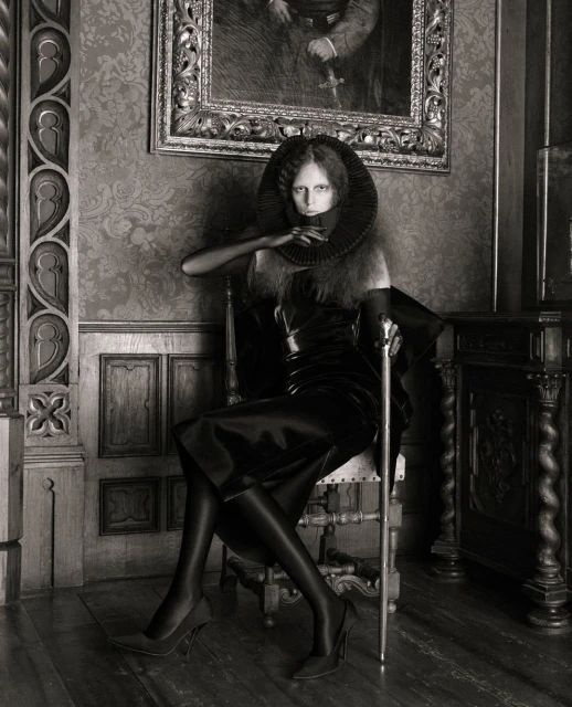 A woman seated on a chair in an elegant interior setting, dressed in a dark outfit with a dramatic collar, and holding a cane.