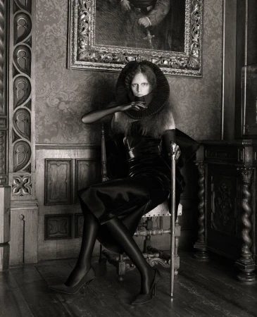 A woman seated on a chair in an elegant interior setting, dressed in a dark outfit with a dramatic collar, and holding a cane.