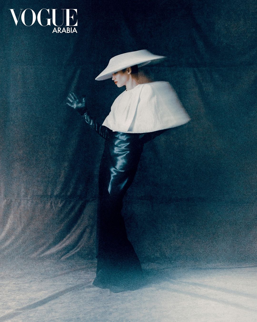 A fashion photograph showcases a slender figure in a black dress and a large white hat against a textured, dark blue backdrop, with subtle lighting.