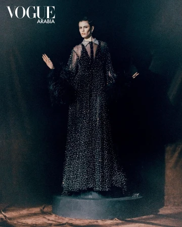 Fashion editorial shot of a model wearing a long black sparkling gown with feather sleeves in front of a dark backdrop.