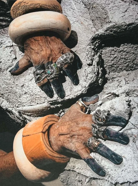 Two hands adorned with rings and bracelets against a textured wall. The jewelry contrasts with the skin's texture and background.