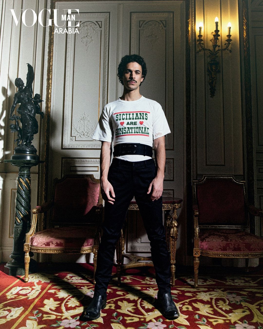 A posed portrait of a man indoors wearing a white t-shirt, black pants, and black boots, with vintage decor and Vogue magazine visible.