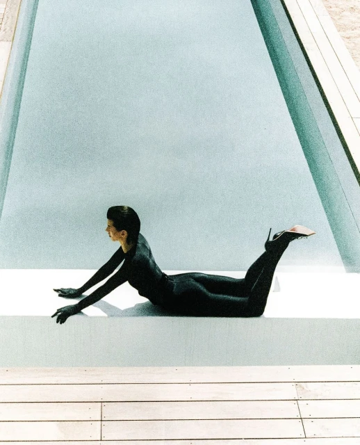 Woman in black catsuit and high heels poses beside a light blue-filled swimming pool, lying on a white platform and deck.