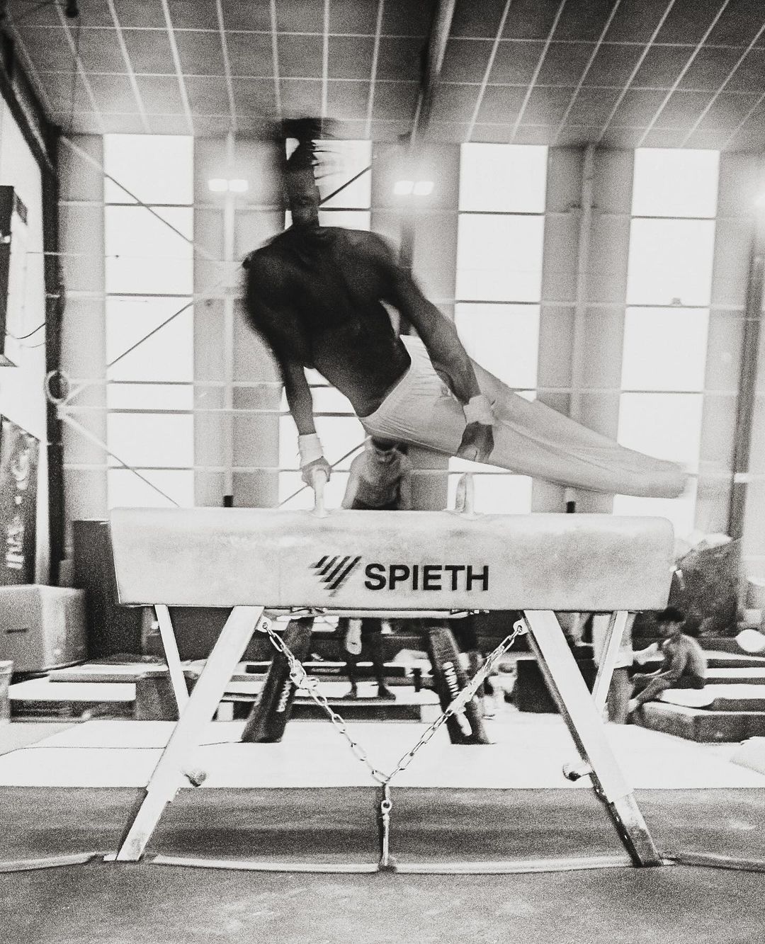 Black and white photo of male gymnast performing on a pommel horse indoors in a gym setting, lit by overhead lighting.