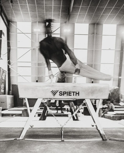 Black and white photo of male gymnast performing on a pommel horse indoors in a gym setting, lit by overhead lighting.