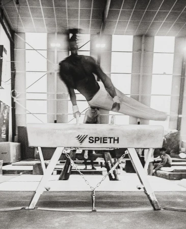 Black and white photo of male gymnast performing on a pommel horse indoors in a gym setting, lit by overhead lighting.