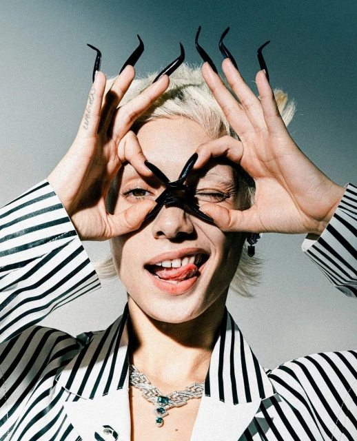 Woman with long black nails makes circle with her hands near her eyes in an indoor portrait with a gray background.