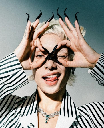 Woman with long black nails makes circle with her hands near her eyes in an indoor portrait with a gray background.